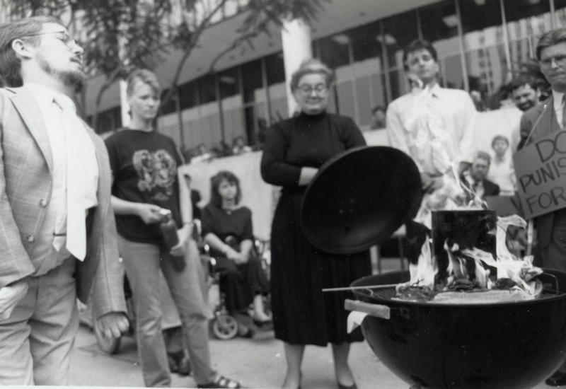 A young Paul Longmore stands in gray plaid suit in front of a weber grill, about to light his book on fire in a protest in front of social security building.